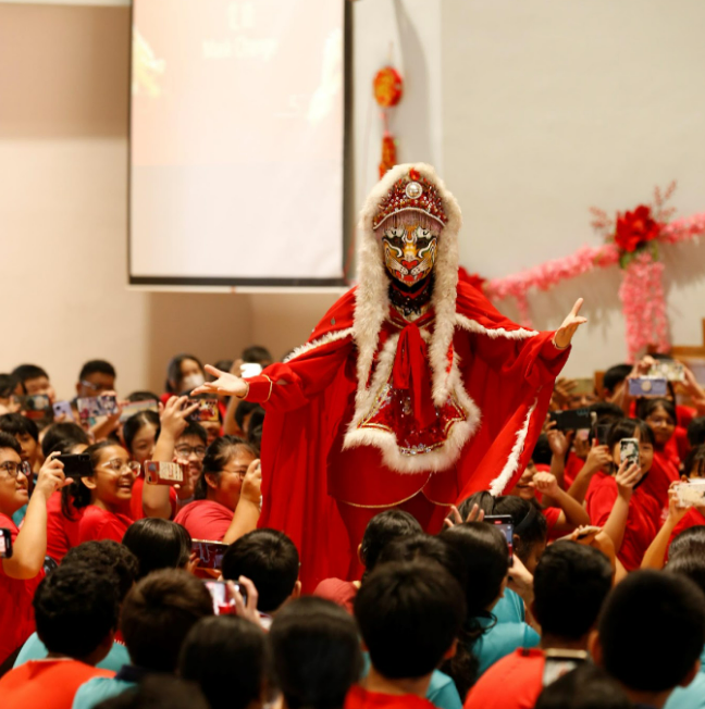A performer in red Chinese traditional costume with ornate headdress walks past a crowd of onlookers taking photos.